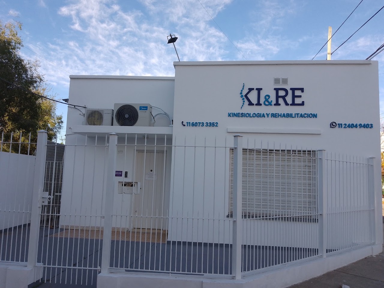 Close-up architectural view of a modern boutique health clinic facade with illuminated sign 'KIRE Kinesiología & Rehabilitación', clean wood and stone textures, glass entrance, welcoming and clinical, sunny daylight in Buenos Aires.
