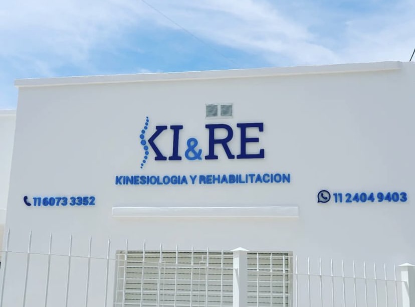 Close-up architectural view of a modern boutique health clinic facade with illuminated sign 'KIRE Kinesiología & Rehabilitación', clean wood and stone textures, glass entrance, welcoming and clinical, sunny daylight in Buenos Aires.