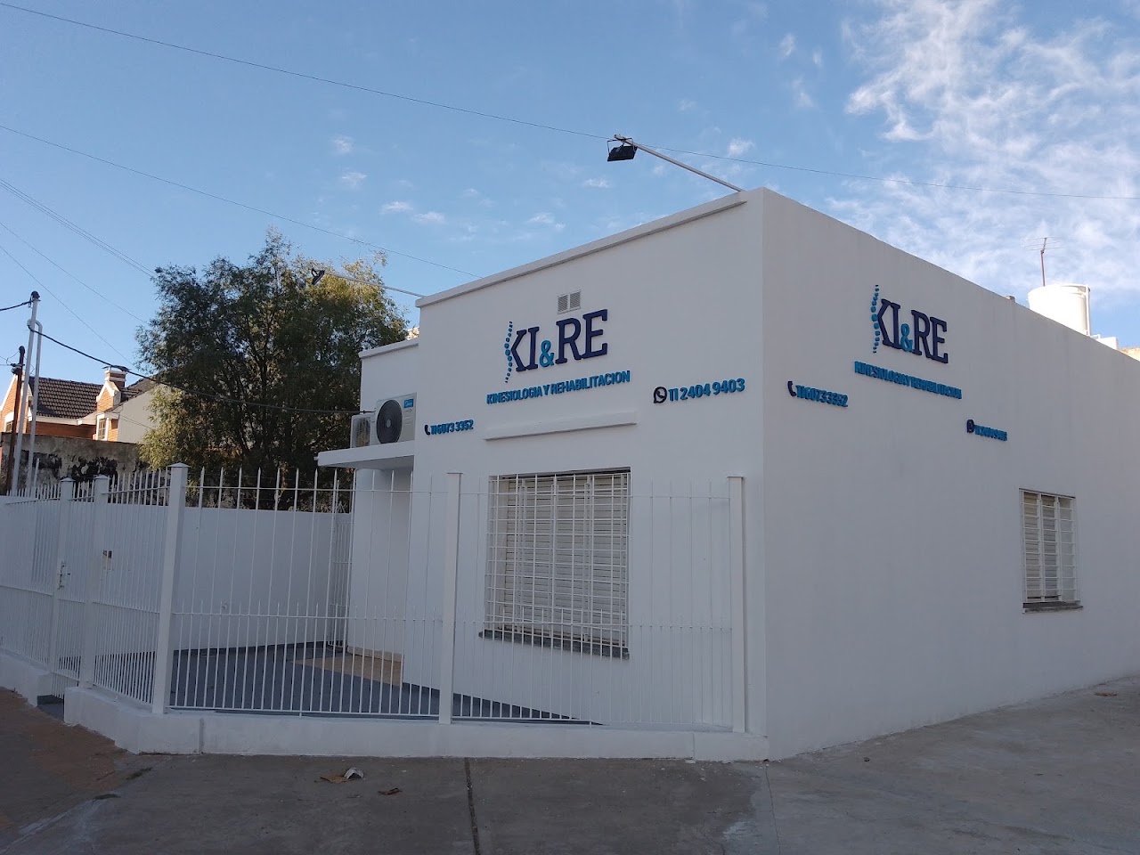 A modern, welcoming facade and entrance of a boutique rehabilitation and physiotherapy clinic named KIRE, clean architectural exterior with glass doors, subtle warm interior lighting, contemporary medical aesthetic in Buenos Aires, landscaped curb, bright daylight, professional architectural photography.
